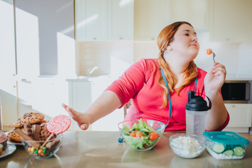 fat young woman in kitchen sitting and eating healthy food. she show stop sign to sweet food and don't look at it. plus size model hold for with tomato on it. healthy food on right side.