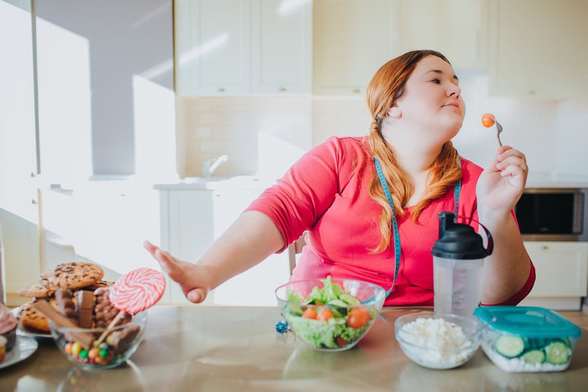 fat young woman in kitchen sitting and eating healthy food. she show stop sign to sweet food and don't look at it. plus size model hold for with tomato on it. healthy food on right side.